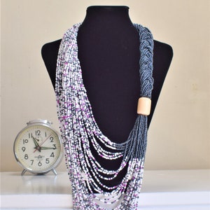 May include: A long, layered necklace made of white, gray, and pink beads. The necklace is draped over a black mannequin and has a wooden bead at the end.