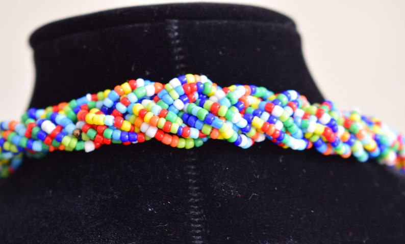 May include: A multi-colored beaded necklace with a braided design. The necklace is made of small, round beads in shades of red, orange, yellow, green, blue, and white.