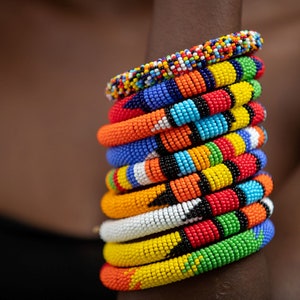 Puede incluir: Una pila de pulseras de cuentas de colores en rojo, naranja, amarillo, verde, azul y blanco. Las pulseras están hechas de pequeñas cuentas redondas y están dispuestas en un patrón.