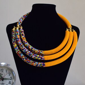 May include: A multi-strand beaded necklace displayed on a black mannequin. The necklace features three strands of bright yellow beads and three strands of colorful, mixed beads. Gold-colored clasps connect the strands.