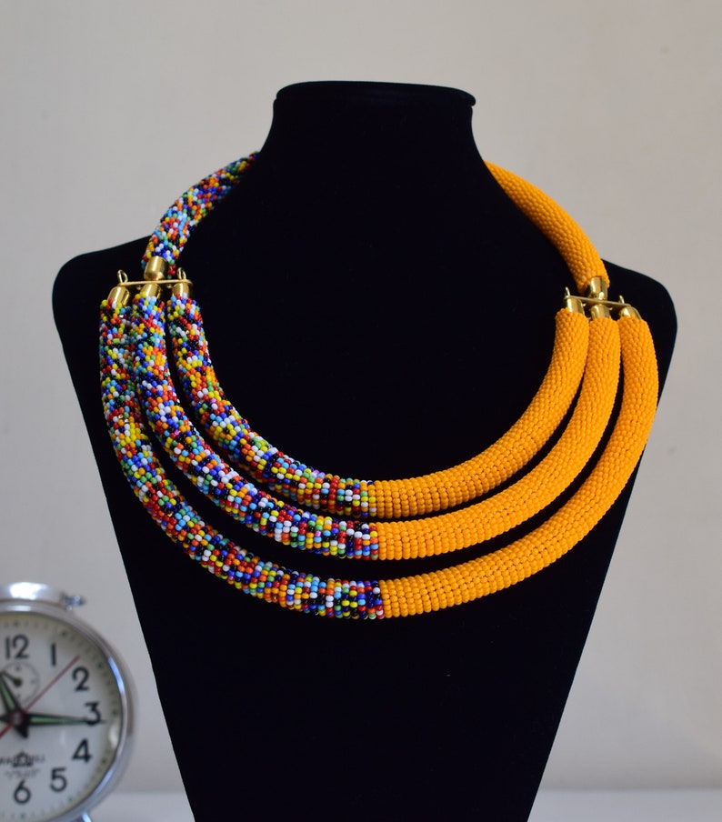 May include: A multi-strand beaded necklace displayed on a black mannequin. The necklace features three strands of bright yellow beads and three strands of colorful, mixed beads. Gold-colored clasps connect the strands.