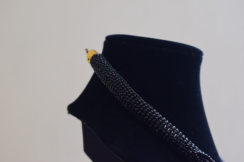 K&ouml;nnte beinhalten: Eine schwarze Perlenkette mit einem goldenen Verschluss auf einem schwarzen Samt-Pr&auml;sentationsst&auml;nder.