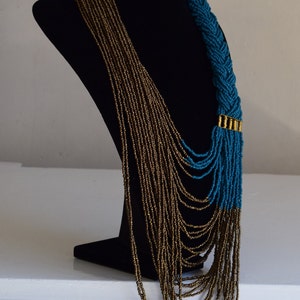 Op de afbeelding: Een lange, gelaagde ketting met een gevlochten gedeelte van teal blauwe kralen en een gouden accent. De ketting is gemaakt van kleine, ronde kralen en wordt getoond op een zwart manekin.