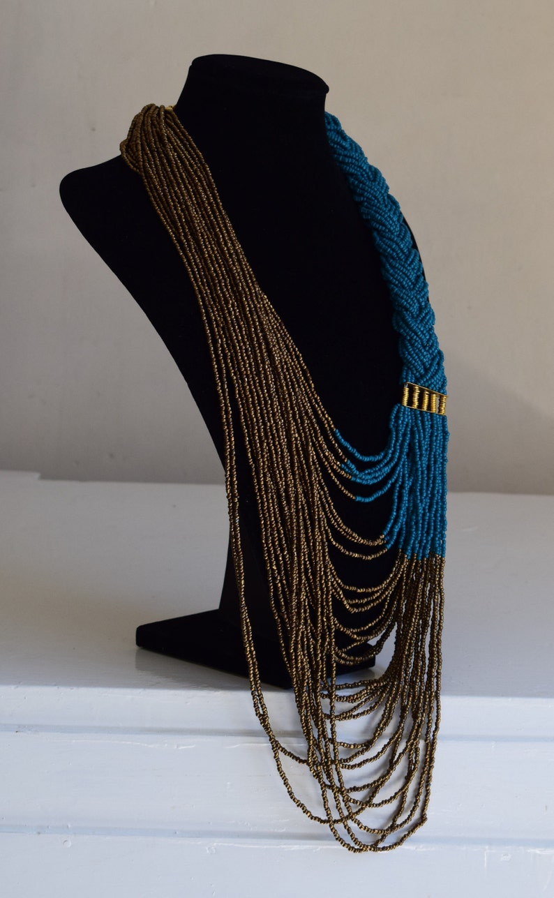 Op de afbeelding: Een lange, gelaagde ketting met een gevlochten gedeelte van teal blauwe kralen en een gouden accent. De ketting is gemaakt van kleine, ronde kralen en wordt getoond op een zwart manekin.
