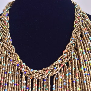 May include: A long, multi-strand necklace made of gold beads with small, colourful beads interspersed throughout. The necklace is braided at the top and hangs in a cascade of beads.
