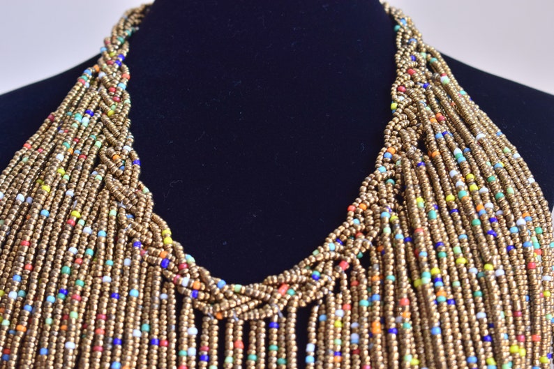May include: A long, multi-strand necklace made of gold beads with small, colourful beads interspersed throughout. The necklace is braided at the top and hangs in a cascade of beads.