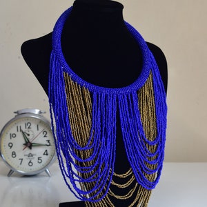 May include: A blue and gold beaded necklace with a long, cascading fringe. The necklace is displayed on a black mannequin bust.