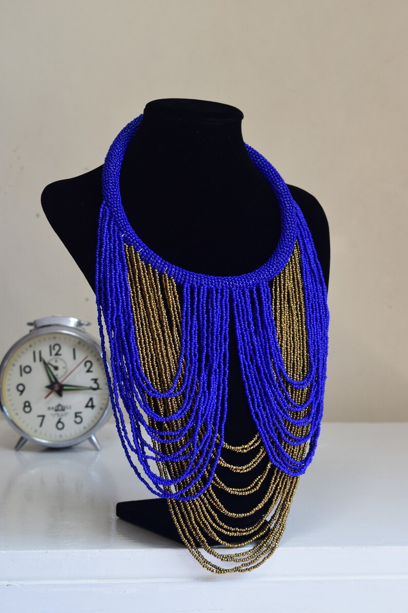 May include: A blue and gold beaded necklace with a long, cascading fringe. The necklace is displayed on a black mannequin bust.
