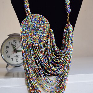 May include: A long, colorful beaded necklace with a large, round, multi-colored beaded pendant. The necklace is made of small, round beads in a variety of colors, including red, blue, green, yellow, and white. The pendant is also made of small, round beads, and it is arranged in a spiral pattern.