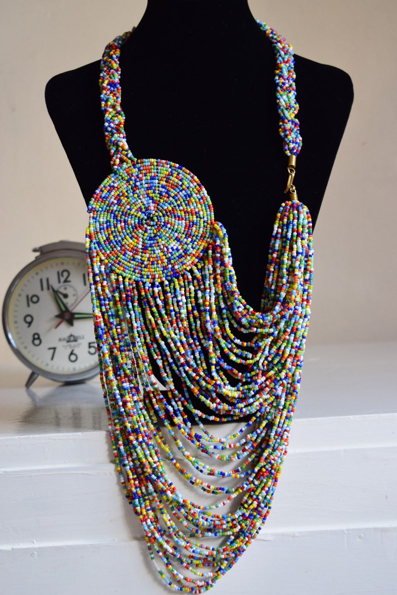 May include: A long, colorful beaded necklace with a large, round, multi-colored beaded pendant. The necklace is made of small, round beads in a variety of colors, including red, blue, green, yellow, and white. The pendant is also made of small, round beads, and it is arranged in a spiral pattern.