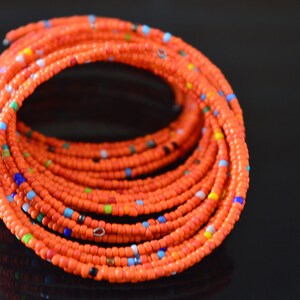 May include: A close-up of a bright orange beaded bracelet with small, colorful beads interspersed throughout. The bracelet is coiled into a circle, showcasing the intricate pattern of the beads.