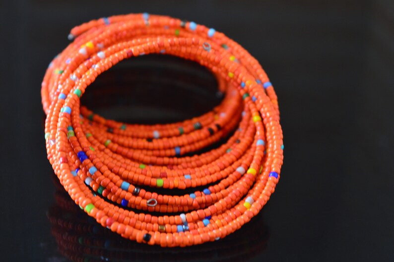 May include: A close-up of a bright orange beaded bracelet with small, colorful beads interspersed throughout. The bracelet is coiled into a circle, showcasing the intricate pattern of the beads.