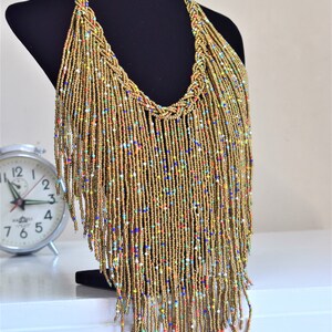 May include: A long gold beaded necklace with colourful beads woven throughout. The necklace has a fringe design and hangs down to the chest.