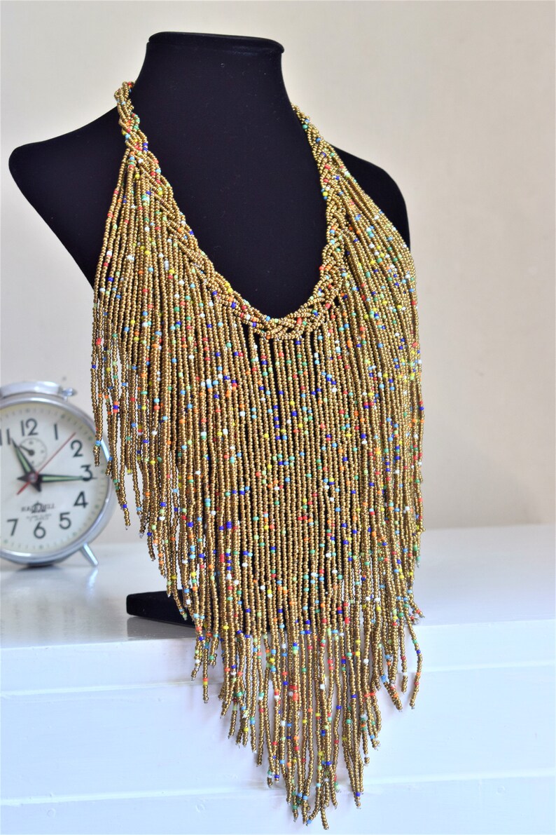 May include: A long gold beaded necklace with colourful beads woven throughout. The necklace has a fringe design and hangs down to the chest.