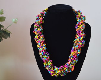 May include: A colorful beaded necklace with a twisted design. The necklace is made of small, round beads in various shades of pink, purple, green, yellow, orange, and brown. The beads are strung on a gold-colored wire.