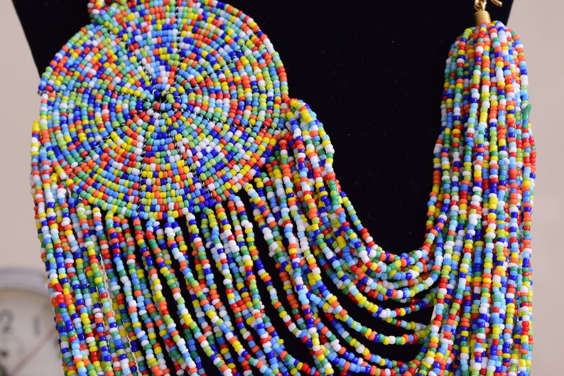 May include: A colorful beaded necklace with a large circular pendant. The necklace is made of small, brightly colored beads in a variety of shades of blue, green, red, yellow, and white. The pendant is a spiral of beads that creates a circular design.