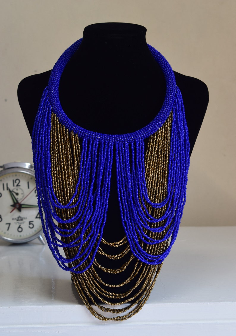 May include: A long, layered necklace made of blue and gold beads. The necklace is displayed on a black mannequin bust.