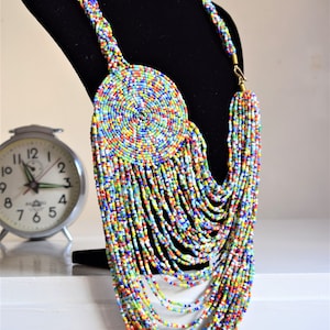 May include: A colorful beaded necklace with a large circular pendant. The necklace is made of small, multi-colored beads and has a long, cascading design. The pendant is also made of beads and has a spiral pattern.