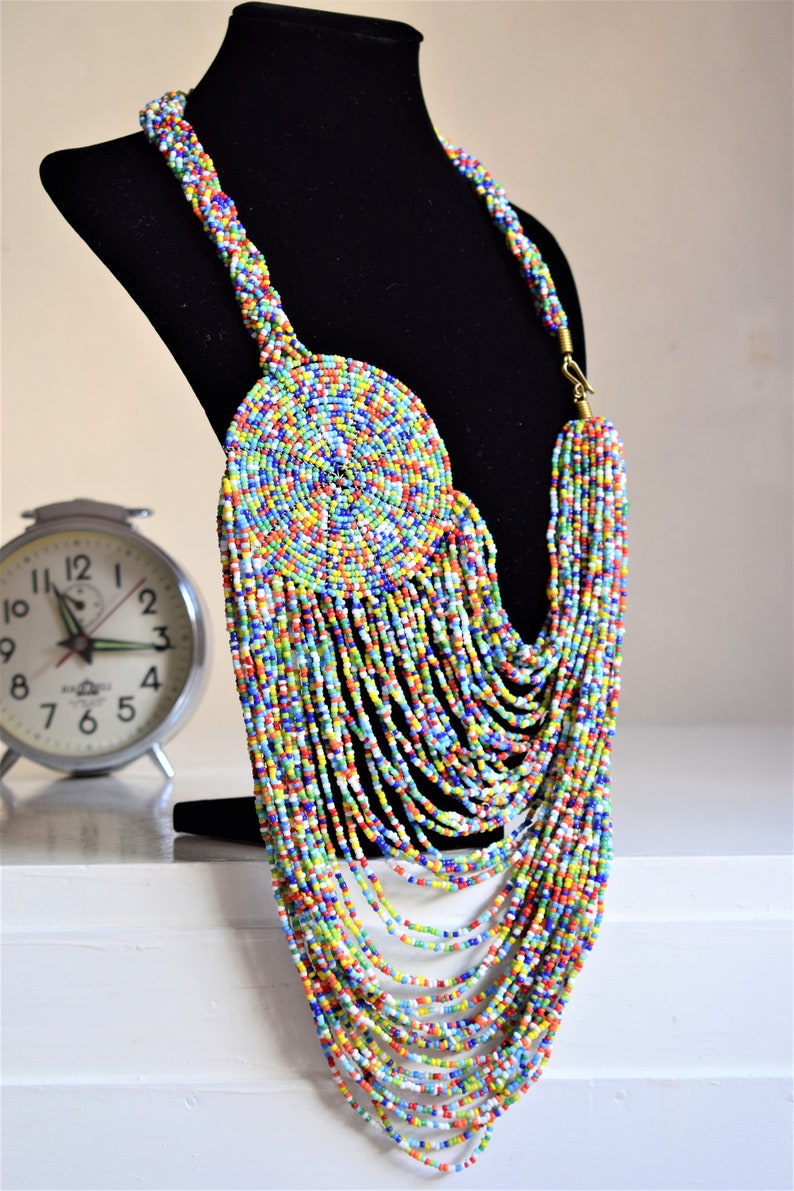 May include: A colorful beaded necklace with a large circular pendant. The necklace is made of small, multi-colored beads and has a long, cascading design. The pendant is also made of beads and has a spiral pattern.