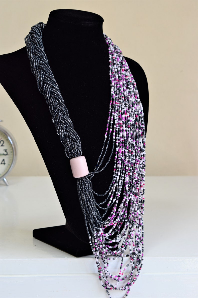 May include: A long, layered necklace made of black, white, and pink beads. The necklace is braided at the top and has a pink bead accent.
