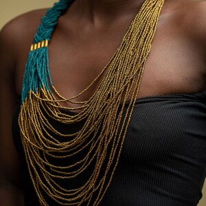 May include: A long, layered necklace with gold and teal beads. The necklace is worn by a person with dark skin.