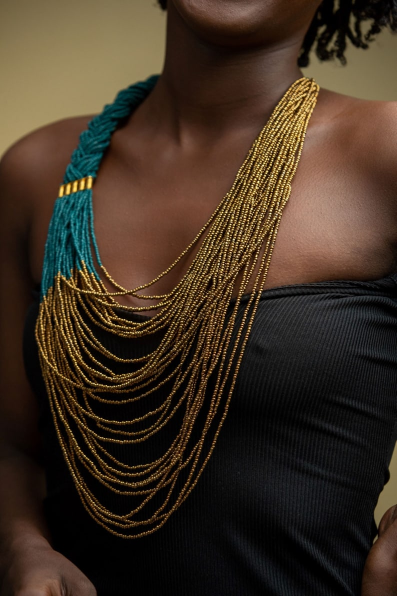 May include: A long, layered necklace with gold and teal beads. The necklace is worn by a person with dark skin.