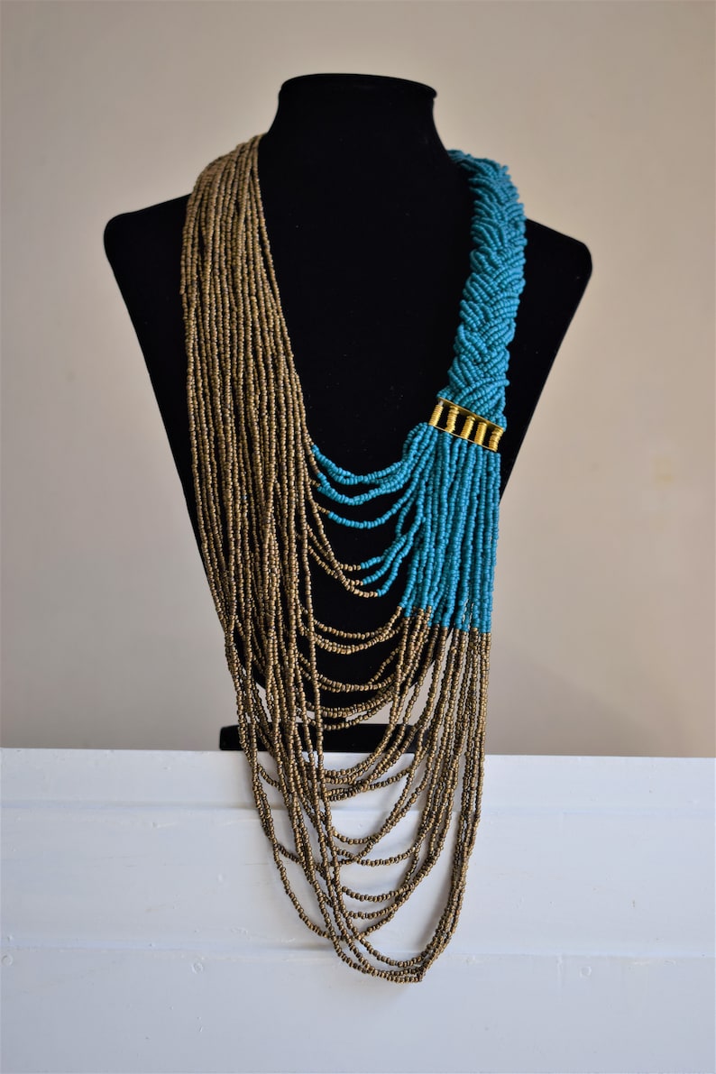 May include: A long, layered necklace with brown and turquoise beads. The necklace is made of multiple strands of beads, with the turquoise beads forming a braided design at the top. The necklace is displayed on a black mannequin.
