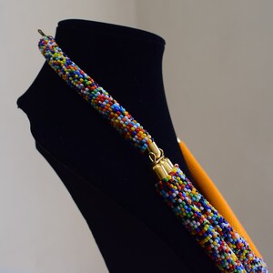 May include: A colorful beaded necklace with a multi-colored pattern of small beads. The necklace has gold-colored clasps and a bright orange backing. The necklace is displayed on a black mannequin.