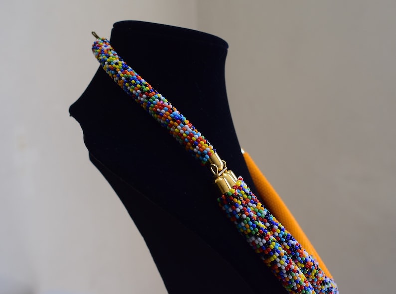 May include: A colorful beaded necklace with a multi-colored pattern of small beads. The necklace has gold-colored clasps and a bright orange backing. The necklace is displayed on a black mannequin.
