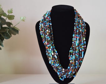 May include: A multi-strand necklace made of small, colorful beads. The beads are in shades of blue, green, orange, red, white, and brown. The necklace is displayed on a black mannequin.