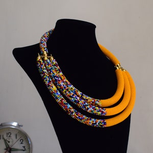 May include: A multi-strand necklace displayed on a black mannequin. The necklace features three strands of bright yellow fabric and three strands of colorful beads. The beads are a mix of red, blue, yellow, and white. The necklace is secured with gold-colored clasps.