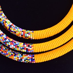 May include: Close-up of three beaded necklaces. Two necklaces are yellow, and the third is a mix of colorful beads. The necklaces are displayed on a black surface, showcasing the vibrant colors and intricate beadwork.