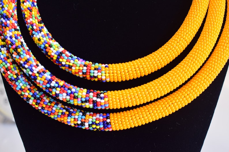 May include: Close-up of three beaded necklaces. Two necklaces are yellow, and the third is a mix of colorful beads. The necklaces are displayed on a black surface, showcasing the vibrant colors and intricate beadwork.