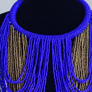 May include: A blue and gold beaded necklace with a fringe design. The necklace is made of small, round beads that are strung together in a long, flowing pattern. The fringe is made of strands of beads that hang down from the bottom of the necklace.