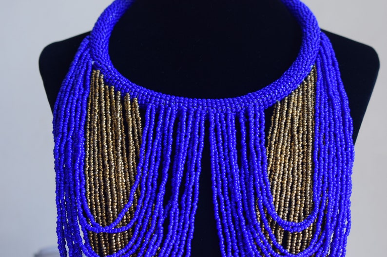 May include: A blue and gold beaded necklace with a fringe design. The necklace is made of small, round beads that are strung together in a long, flowing pattern. The fringe is made of strands of beads that hang down from the bottom of the necklace.