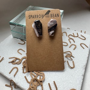 May include: A pair of black and white coffin-shaped earrings with gold flecks. The earrings are displayed on a brown card with the text "Sparrow and Bean".