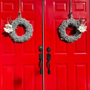 May include: Two red double doors with black door handles and two gray wreaths with white flowers hanging on them.