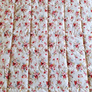 May include: A white quilted fabric with a floral pattern featuring pink, red, and blue flowers. The fabric has a repeating pattern of small floral bouquets.