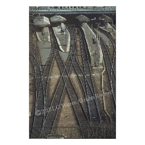 May include: A high-angle view of a train track junction with multiple sets of tracks crossing each other. The tracks are made of metal and are surrounded by gravel. There are several buildings in the background, including a train station.