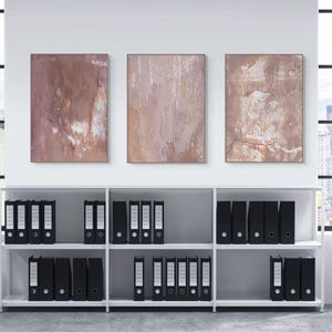 Pode incluir: Três pinturas abstratas em tons de castanho e branco penduradas acima de uma estante branca cheia de pastas pretas. As pinturas têm uma aparência texturizada. A sala tem janelas grandes com vista para a cidade.