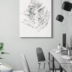Può includere: Stampa su tela incorniciata con un'impressione dettagliata di una foglia di felce in tonalità grigie e bianche. L'opera d'arte è appesa a una parete grigio chiaro in un ambiente ufficio moderno, completata da sedie grigie e una scrivania.
