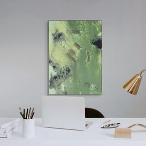May include: A rectangular wall art piece featuring an abstract composition of blended sage green, olive, and dark charcoal tones. The artwork is displayed in a thin frame and hangs above a white desk with a laptop, a lamp, and a stack of books.