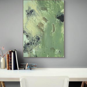Puede incluir: Impresión artística abstracta con orientación vertical. La obra presenta una mezcla de tonos verdes, amarillos y negros, creando un diseño texturizado y fluido. La pieza está enmarcada y montada en una pared gris, encima de un escritorio blanco.