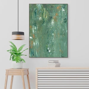 May include: Abstract green and white painting in a black frame. The artwork features textured brushstrokes with hints of orange. The painting is displayed above a wooden cabinet with a plant and a lamp.