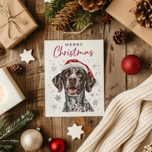 以下が含まれることがあります： 赤いサンタの帽子をかぶった茶色と白の犬が描かれたクリスマスカード。カードは白い背景に雪の結晶が描かれており、ピンク色の「メリークリスマス」の文字が書かれている。