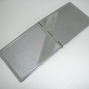 May include: A silver metal stamp case with a hinged lid. The case has a textured surface and the word "Stamps" is engraved on the lid.