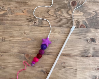 May include: A wooden stick with a string attached to a star-shaped felt toy. The string has a series of felt balls in shades of pink, purple, and red.