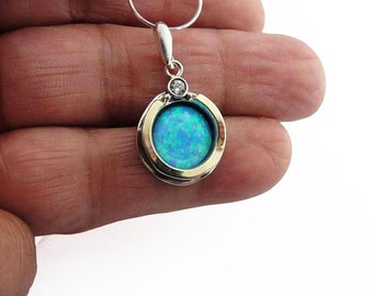 May include: A round pendant with a silver chain. The pendant features a gold-colored bezel surrounding a vibrant blue opal stone. A small, clear gemstone is set above the opal. The pendant is held in a hand.