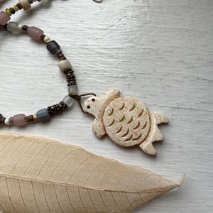 May include: A beaded necklace with a turtle pendant. The necklace features a variety of beads in shades of gray, pink, and brown. The turtle pendant is a light beige color with carved details. A dried leaf is also present.