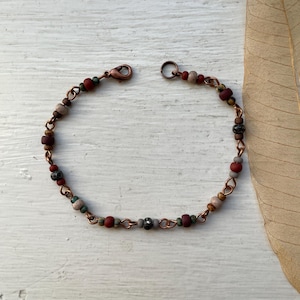 May include: A copper chain bracelet featuring a series of small, multi-coloured beads in shades of terracotta, sage green, grey, and tan. The bracelet is secured with a copper lobster claw clasp and includes a circular jump ring for closure.